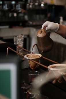 A barista pours water from a rustic, metal kettle into a coffee dripper containing ground coffee, and the brewed coffee flows into a glass carafe below. The setting appears to be a cafe with a warm ambiance.