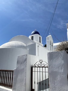 A whitewashed building with a blue domed roof and cross on top under a clear blue sky. The architecture is typical of Greek Orthodox churches, featuring simple, geometric shapes and minimal ornamentation. A wrought iron gate with decorative scrollwork stands in the foreground.