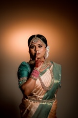 A woman dressed in traditional attire with a silk saree in shades of green and gold, adorned with intricate jewelry, including a necklace, earrings, and a headpiece. Her hand is decorated with henna and she gestures with a finger to her lips suggesting silence. The background is softly lit with a warm glow, highlighting her poised and graceful stance.