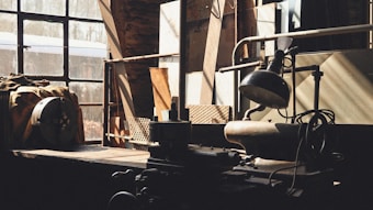 A dimly lit industrial workshop with a large window allowing beams of natural light to filter through. The room contains various machinery and tools, including a lathe, a lamp, and storage shelves. The setting appears aged and used, with dust and signs of wear on surfaces.