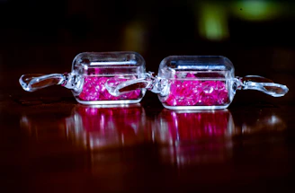 Two small, rectangular, transparent containers with handles sit side by side on a reflective, dark surface. Each container is filled with small, vibrant pink crystals. The overall atmosphere is highlighted by reflections and a soft focus blending with a dark background.