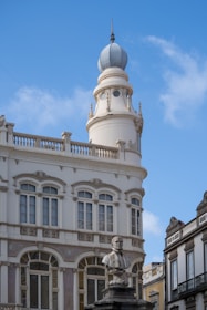 A historical building with ornate architecture featuring tall arched windows and decorative details. At the top of the building, there is a round tower with a domed roof and decorative balustrades. In front of the building, a stone bust of a man is mounted on a pedestal, adding to the classical aesthetic.