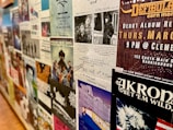 A wall covered with various posters and flyers for music events and performances. Most of the posters feature bands, details of concert dates and locations, and artistic designs. The wall has a collage effect with overlapping elements creating a busy and colorful atmosphere.