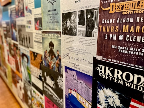 A wall covered with various posters and flyers for music events and performances. Most of the posters feature bands, details of concert dates and locations, and artistic designs. The wall has a collage effect with overlapping elements creating a busy and colorful atmosphere.