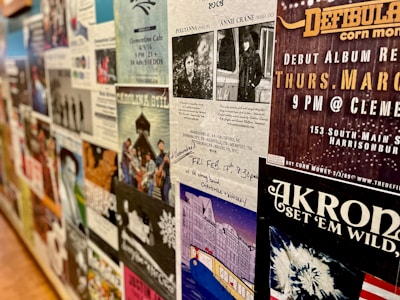 A wall covered with various posters and flyers for music events and performances. Most of the posters feature bands, details of concert dates and locations, and artistic designs. The wall has a collage effect with overlapping elements creating a busy and colorful atmosphere.