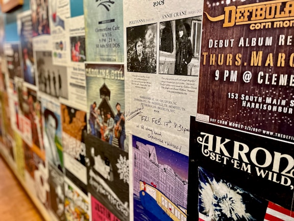 A wall covered with various posters and flyers for music events and performances. Most of the posters feature bands, details of concert dates and locations, and artistic designs. The wall has a collage effect with overlapping elements creating a busy and colorful atmosphere.