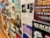 A wall covered with various posters and flyers for music events and performances. Most of the posters feature bands, details of concert dates and locations, and artistic designs. The wall has a collage effect with overlapping elements creating a busy and colorful atmosphere.