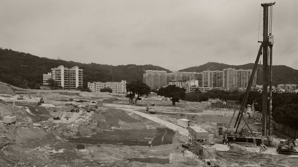 A large construction site featuring machinery and equipment situated in a wide, open landscape. In the background, modern high-rise residential buildings are visible against a hilly landscape.