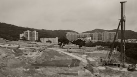 A large construction site featuring machinery and equipment situated in a wide, open landscape. In the background, modern high-rise residential buildings are visible against a hilly landscape.
