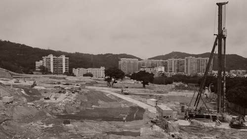 A large construction site featuring machinery and equipment situated in a wide, open landscape. In the background, modern high-rise residential buildings are visible against a hilly landscape.