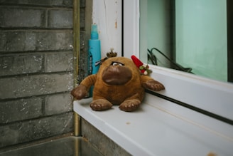 A plush toy resembling a cartoonish animal with large feet and a big nose sits on a windowsill. Next to it is a small, colorful decoration. The setting includes a blue bottle and a textured brick wall, creating a cozy and somewhat whimsical atmosphere.