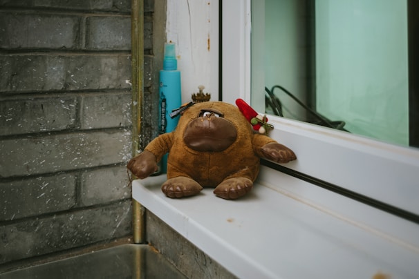 A plush toy resembling a cartoonish animal with large feet and a big nose sits on a windowsill. Next to it is a small, colorful decoration. The setting includes a blue bottle and a textured brick wall, creating a cozy and somewhat whimsical atmosphere.