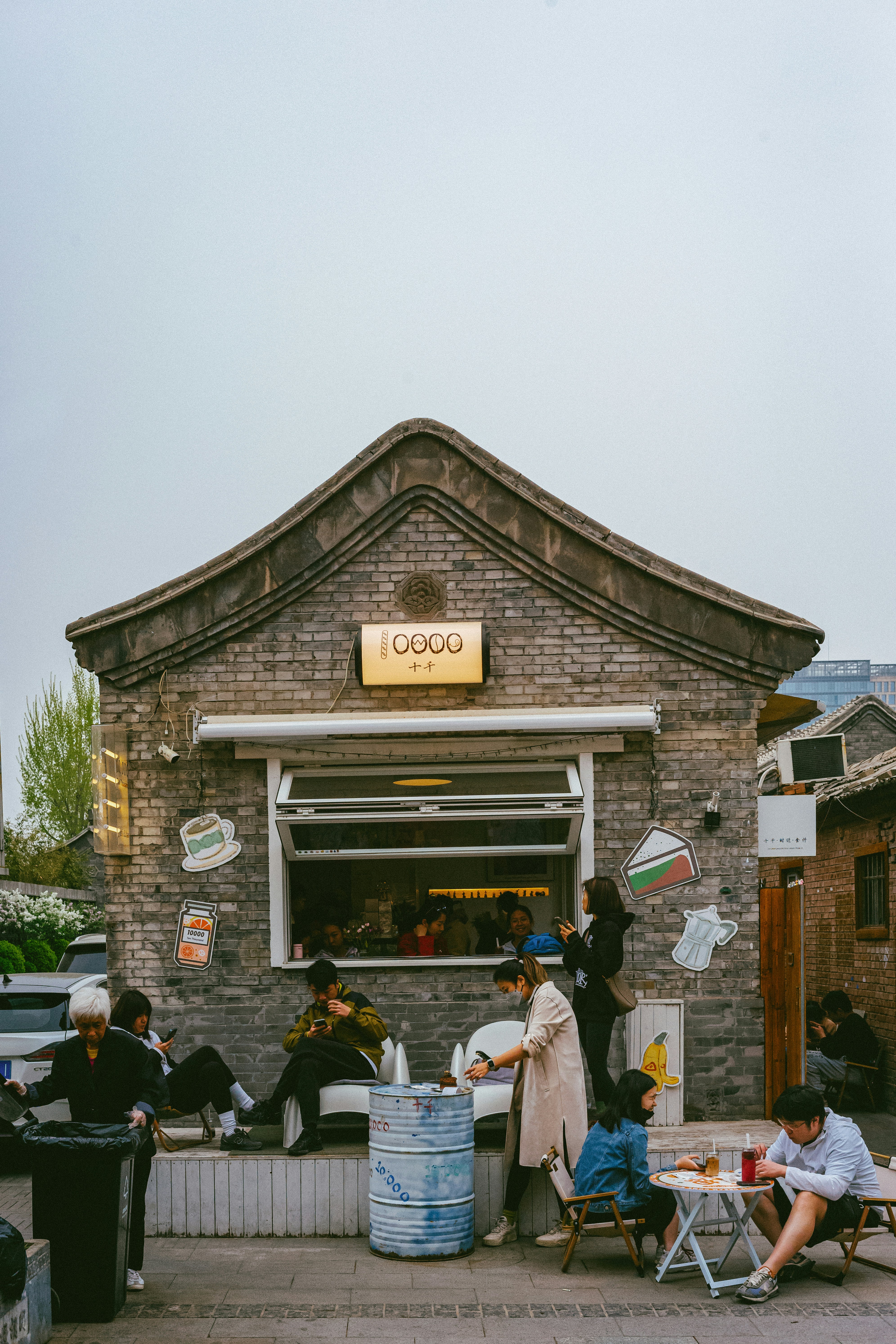 A quaint cafe with a traditional brick facade features people seated outside, some using their phones. There are cartoonish stickers of coffee cups and other items on the walls. The atmosphere is casual and relaxed.