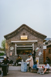 A quaint cafe with a traditional brick facade features people seated outside, some using their phones. There are cartoonish stickers of coffee cups and other items on the walls. The atmosphere is casual and relaxed.