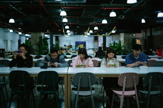 A group of five people sitting at a long table in a modern, well-lit cafeteria. They are all engaged with their smartphones and appear to be focused. Various plants and decorations can be seen in the background, giving a relaxed and contemporary atmosphere.