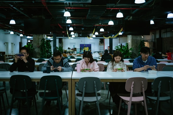 A group of five people sitting at a long table in a modern, well-lit cafeteria. They are all engaged with their smartphones and appear to be focused. Various plants and decorations can be seen in the background, giving a relaxed and contemporary atmosphere.