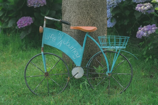 An old-fashioned turquoise bicycle with a large front card that has 'Life is a Journey' written on it is leaning against a tree. The bicycle has a brown leather seat and a wire basket attached to its back. It is placed on a lush green grassy lawn with blooming purple and blue hydrangea flowers in the background.
