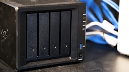 A black network-attached storage (NAS) device with four drive bays is placed on a black table. Blue and white cables are visible in the background.