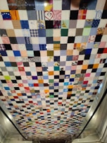 A patchwork quilt comprised of numerous small square pieces of fabric in various colors and patterns. The quilt hangs vertically, showcasing a mix of bright and muted tones with some squares featuring recognizable prints and designs such as hearts and flowers.