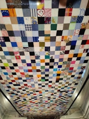 A patchwork quilt comprised of numerous small square pieces of fabric in various colors and patterns. The quilt hangs vertically, showcasing a mix of bright and muted tones with some squares featuring recognizable prints and designs such as hearts and flowers.