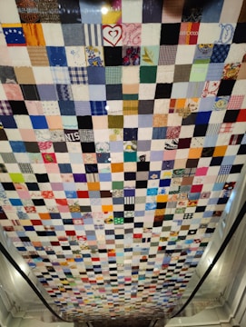 A patchwork quilt comprised of numerous small square pieces of fabric in various colors and patterns. The quilt hangs vertically, showcasing a mix of bright and muted tones with some squares featuring recognizable prints and designs such as hearts and flowers.