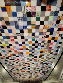 A patchwork quilt comprised of numerous small square pieces of fabric in various colors and patterns. The quilt hangs vertically, showcasing a mix of bright and muted tones with some squares featuring recognizable prints and designs such as hearts and flowers.