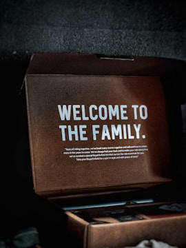 A cardboard box with a message inside that reads 'WELCOME TO THE FAMILY.' Additional smaller text provides a warm welcome, mentioning years of riding together and a curated Royal Enfield kit. The box appears partially open with some visible contents, though they are indistinct.