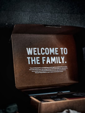 A cardboard box with a message inside that reads 'WELCOME TO THE FAMILY.' Additional smaller text provides a warm welcome, mentioning years of riding together and a curated Royal Enfield kit. The box appears partially open with some visible contents, though they are indistinct.
