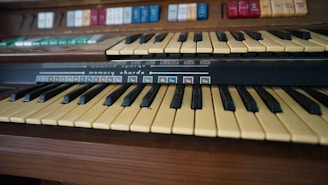 A close-up view of an electronic organ keyboard with two rows of keys and a panel of labeled buttons above. The keys are black and yellowish-white, and the buttons are in various colors with text labels.