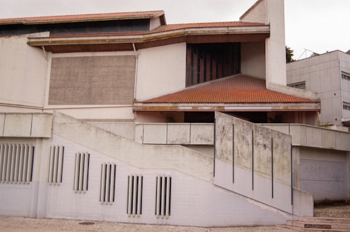 The image depicts a modern architectural building with a combination of geometric shapes and a sloped roof. The structure features a facade with vertical slats and a concrete wall that shows signs of weathering. There are multiple levels and sections, including a prominent stairway and a mix of open and enclosed spaces.