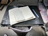 An open notebook filled with handwritten notes is placed on a black makeshift table, which appears to be resting on a trash can. A blue pen lies next to the notebook. The setting seems to be in a garage or workshop, with storage boxes and wooden planks visible in the background.