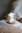 A sculpted candle in the shape of a woman's torso is placed on a wooden base. The background includes soft-focus dried flowers and a rolled twine. The setting is calm with a neutral color palette, and there is a gentle natural light.