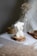A sculpted candle in the shape of a woman's torso is placed on a wooden base. The background includes soft-focus dried flowers and a rolled twine. The setting is calm with a neutral color palette, and there is a gentle natural light.