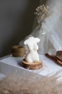 A sculpted candle in the shape of a woman's torso is placed on a wooden base. The background includes soft-focus dried flowers and a rolled twine. The setting is calm with a neutral color palette, and there is a gentle natural light.