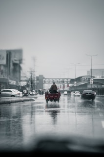 A rainy street scene with wet roads reflects vehicles driving on a thoroughfare. In the center, a small three-wheeled vehicle with a person sitting on its back moves along the road. Surrounding the main street, there are several cars and city buildings blurred in the background under a gray overcast sky.