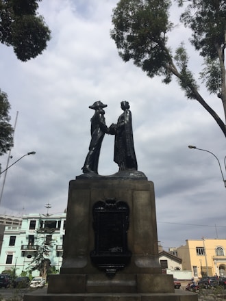 A statue of two figures in historical attire is prominently displayed on a pedestal. They are shaking hands in a setting surrounded by trees and urban buildings under a cloudy sky.