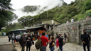 A group of people gathered near a stone wall and a building at the base of a mountain. The area is surrounded by lush greenery, and mist or clouds are visible in the mountainous background. Many of the individuals are wearing masks and carrying backpacks, suggesting a travel or tourism setting. A bus is parked nearby.