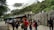 A group of people gathered near a stone wall and a building at the base of a mountain. The area is surrounded by lush greenery, and mist or clouds are visible in the mountainous background. Many of the individuals are wearing masks and carrying backpacks, suggesting a travel or tourism setting. A bus is parked nearby.