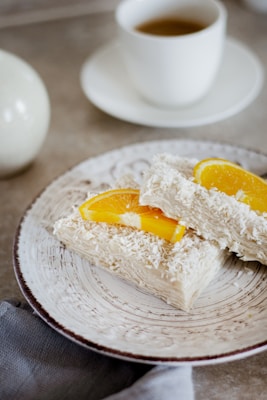 A rustic plate contains two pieces of dessert topped with shredded coconut and garnished with fresh orange slices. In the background, there is a cup of tea or coffee on a saucer, suggesting a cozy, relaxed setting. The plate and the background surface have a neutral, earthy tone, enhancing the simplicity and elegance of the presentation.