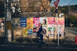 A woman in traditional Japanese attire walks by a wall covered with various colorful posters featuring faces and text, likely for political campaigns, under a stop sign.