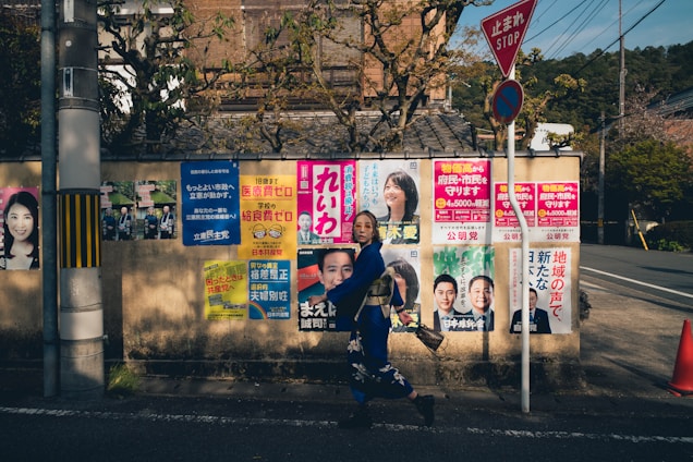 A woman in traditional Japanese attire walks by a wall covered with various colorful posters featuring faces and text, likely for political campaigns, under a stop sign.