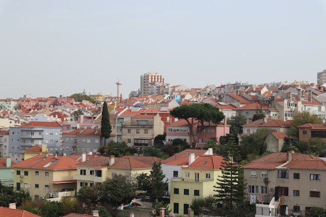 A residential area featuring an array of buildings with red-tiled roofs, positioned on a gently rising hill. The architecture varies with some structures painted in pastel colors like blue and pink, along with neutral tones. In the foreground, there is lush greenery with trees and bushes, creating a serene contrast to the urban setup. A construction crane is visible in the distance, indicating ongoing development. The skyline is dominated by a pale sky, with visually striking taller buildings in the backdrop.