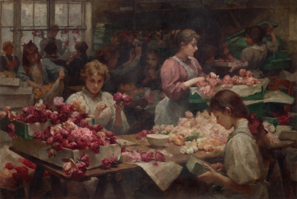Several young women are engaged in arranging and packing roses within a dimly lit workshop. Roses, predominantly in shades of red and pink, cover the tables and are stacked in boxes. The setting suggests a sense of industrious activity, with each individual focused on handling the flowers.