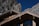 A wooden chalet roof with intricate detailing is set against a backdrop of rugged, rocky mountains under a clear blue sky. The sunlight casts shadows, enhancing the textures of the wood and the rocks.