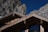 A wooden chalet roof with intricate detailing is set against a backdrop of rugged, rocky mountains under a clear blue sky. The sunlight casts shadows, enhancing the textures of the wood and the rocks.