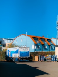 A blue truck with white text is parked in front of a light blue building with orange roofs. Stacks of beverage crates are arranged beside the truck and building. The sky is clear and blue.
