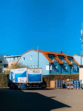 A blue truck with white text is parked in front of a light blue building with orange roofs. Stacks of beverage crates are arranged beside the truck and building. The sky is clear and blue.