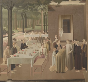 A serene outdoor scene depicting multiple people gathered around long tables set with white tablecloths. The setting appears to be a wooded area with tall trees in the background. People are socializing, some sitting while others are standing, engaged in conversation. A few individuals are seen serving or holding food items like watermelon.