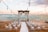An outdoor wedding setup on a wooden deck by the ocean features white chairs arranged in rows facing an altar adorned with flowing white drapes and floral decorations. String lights are hung above, and the ocean provides a serene backdrop with a pastel sky.
