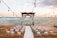 An outdoor wedding setup on a wooden deck by the ocean features white chairs arranged in rows facing an altar adorned with flowing white drapes and floral decorations. String lights are hung above, and the ocean provides a serene backdrop with a pastel sky.